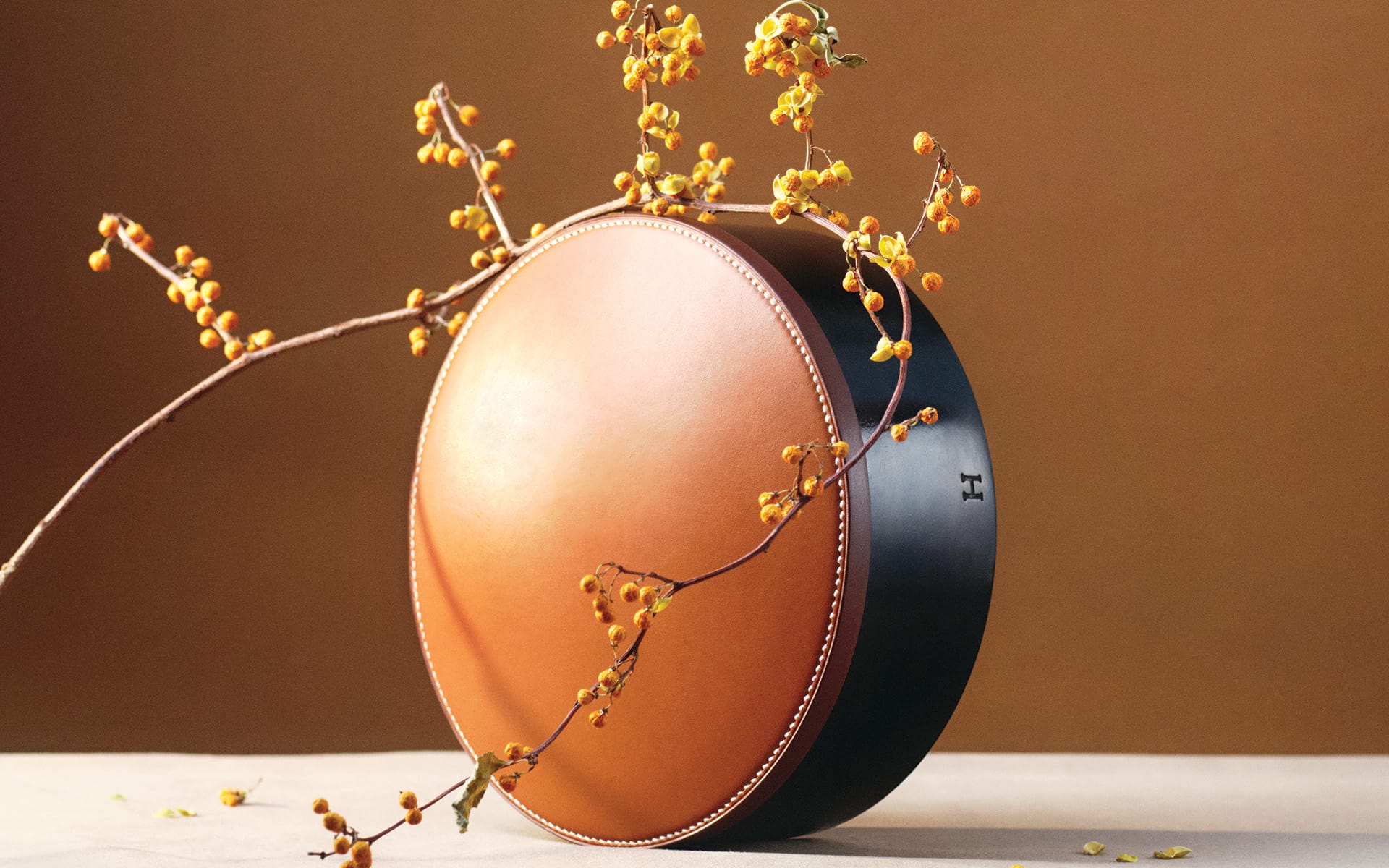 Round leather box with decorative stitches and small branches with yellow berries on a neutral background.