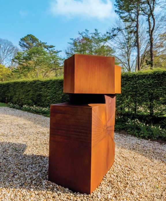 Rust-colored geometric sculpture on gravel path, surrounded by lush greenery and tall trees under a clear blue sky.