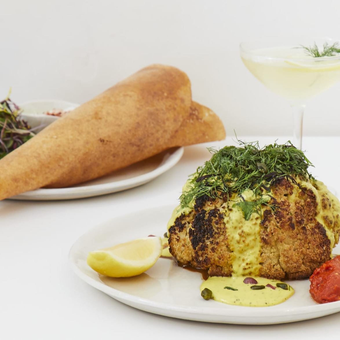 Roasted cauliflower with herb garnish, lemon wedge, and side dishes on a white plate with a cocktail in the background