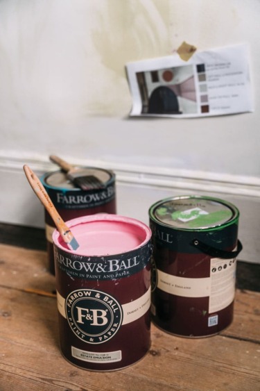 Paint cans and brushes on a wooden floor in front of a wall with a color chart.