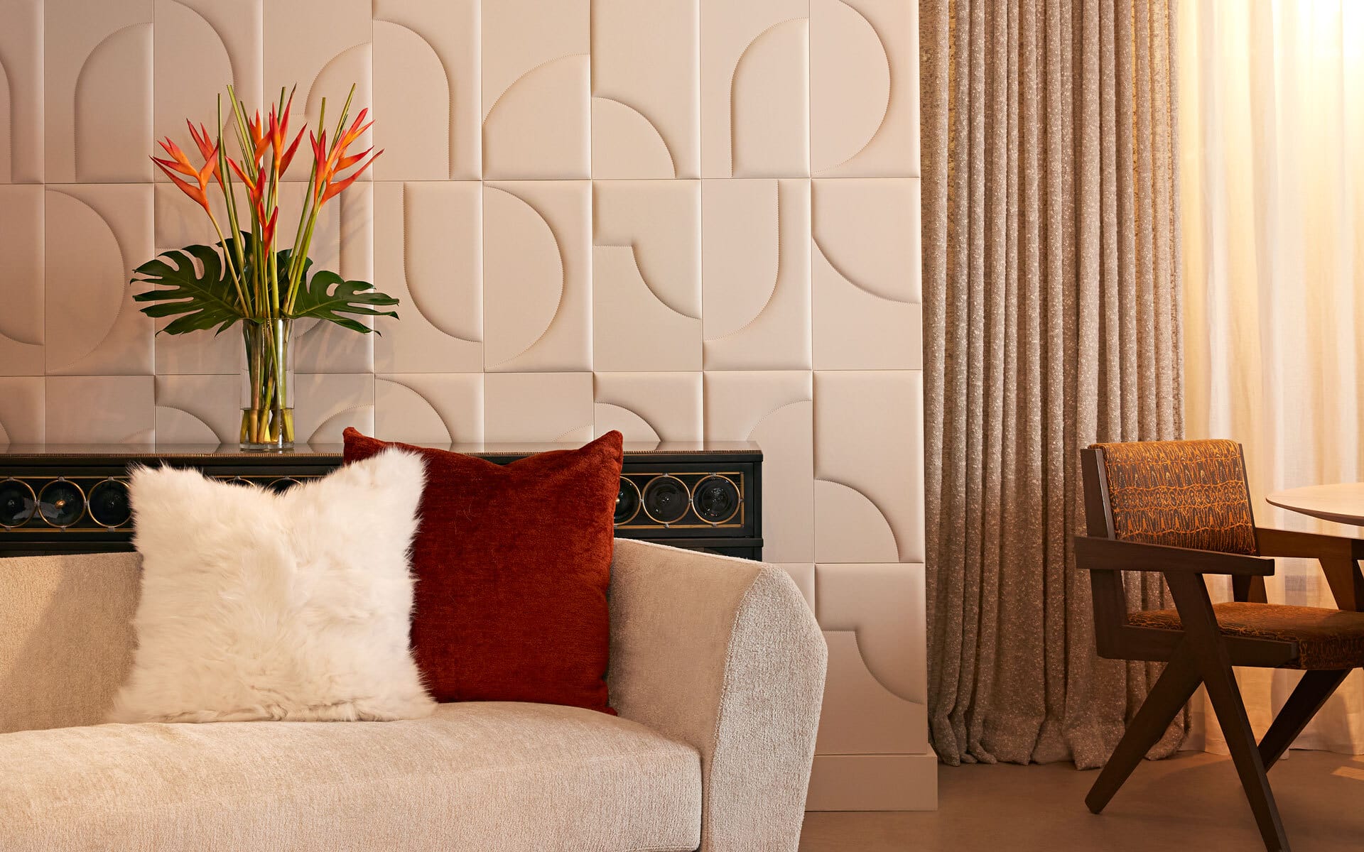 Modern living room with textured wall, beige sofa, fluffy white and red cushions, floral arrangement, and wooden chair.