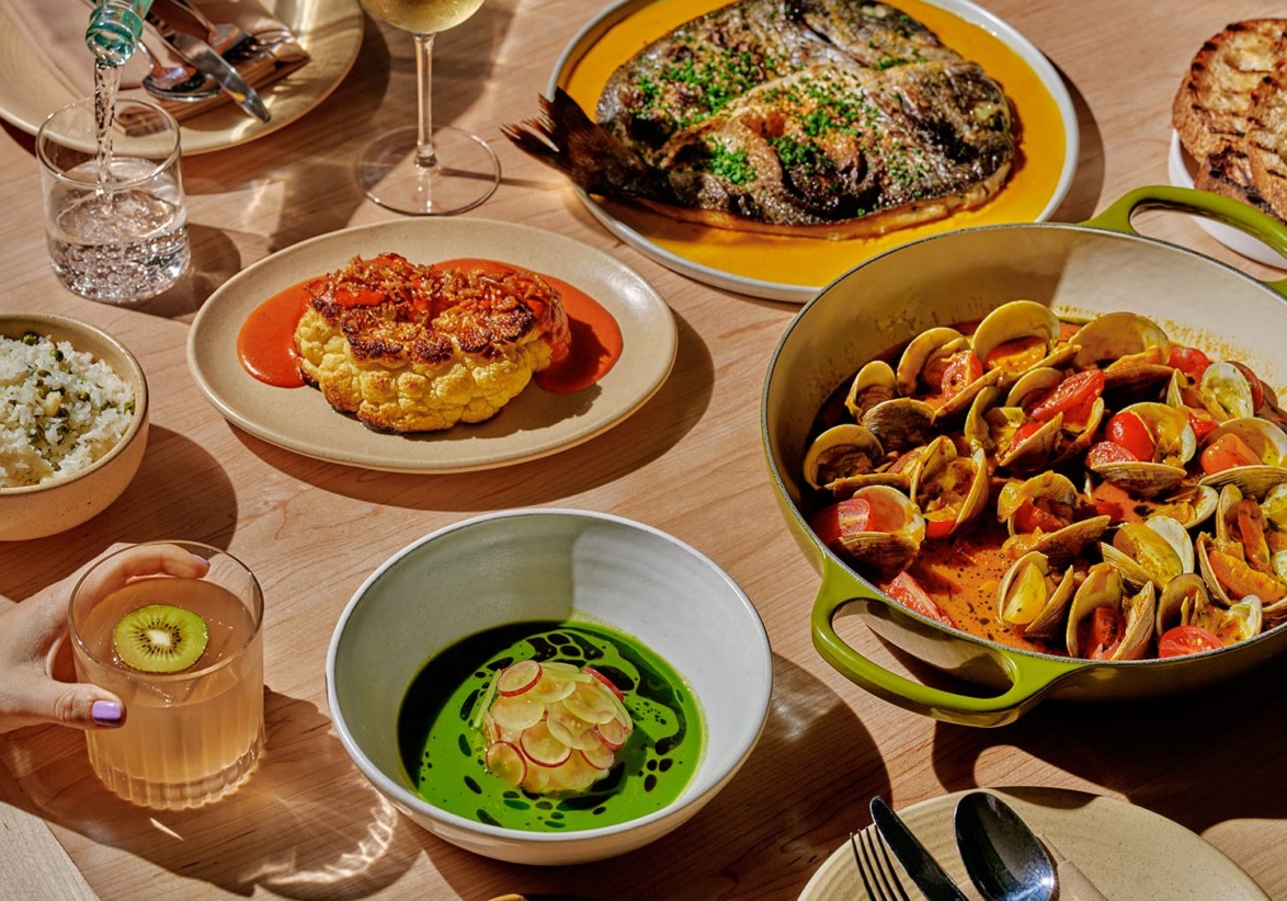 A variety of dishes on a wooden table, including seafood stew, grilled fish, cauliflower dish, and a kiwi drink.