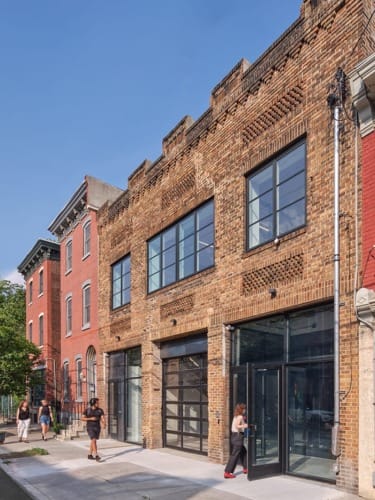 Historic brick building with large windows and people walking on the sidewalk under a clear blue sky.