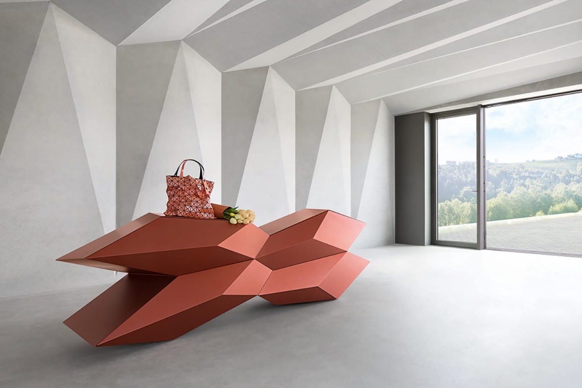 Modern geometric red-orange table with a patterned handbag and flowers in a minimalist room with geometric walls and large window.