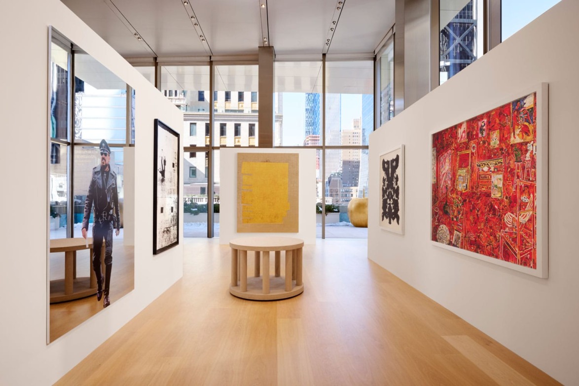 Art gallery with multiple colorful paintings on white walls, large windows, and a wooden table in the center.