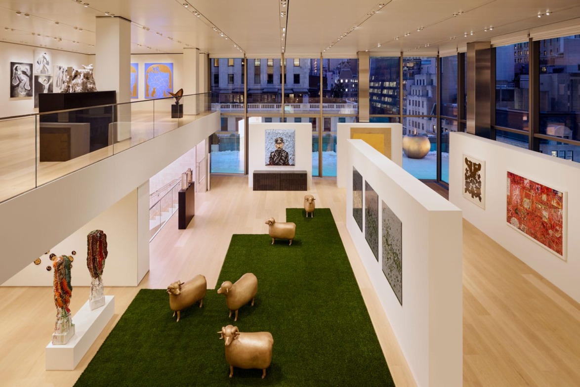 Contemporary art gallery interior with sculptures of golden sheep, paintings on white walls, and large windows revealing cityscape.