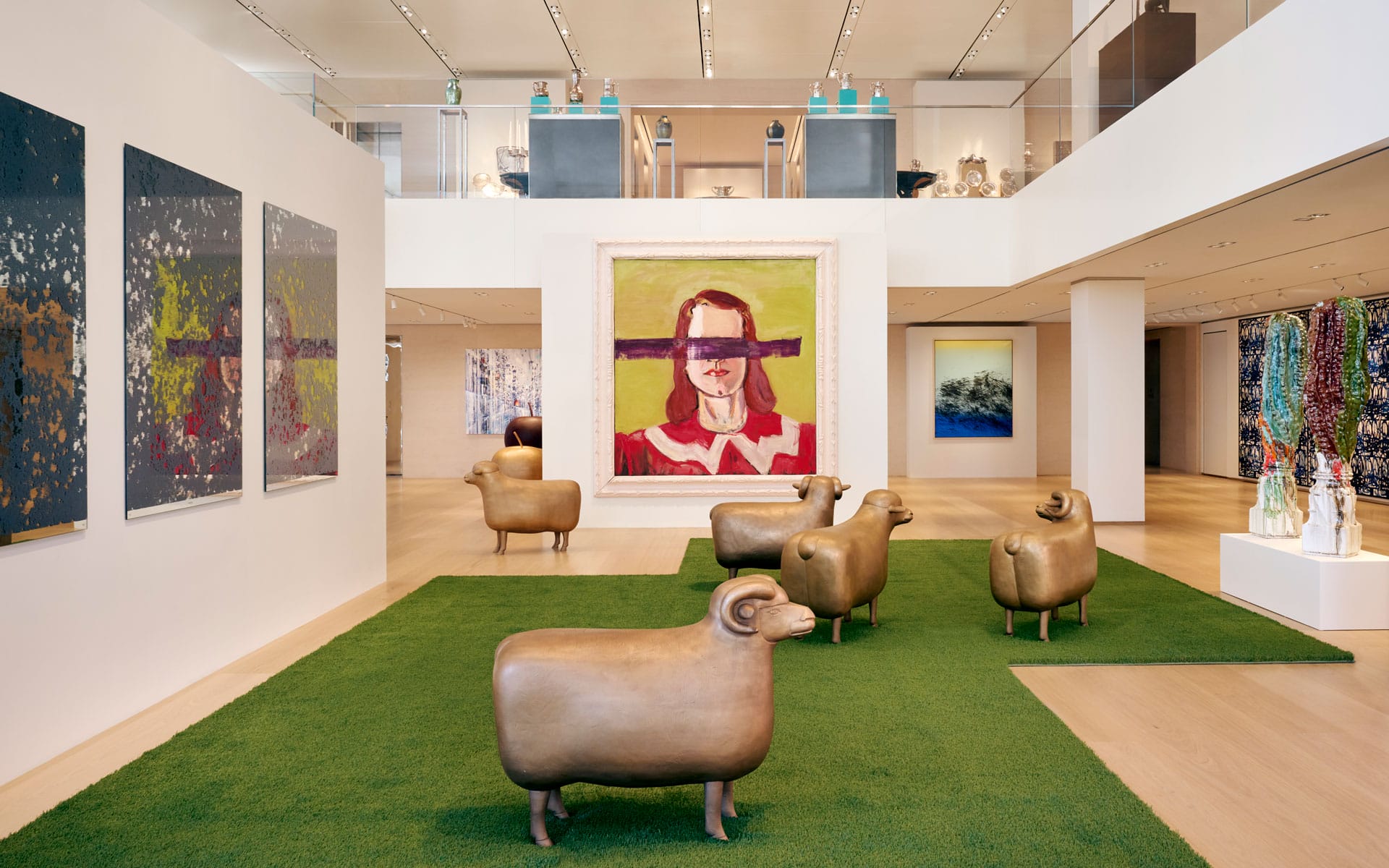 Gallery interior with modern art paintings on walls, bronze sheep sculptures on green rug, wooden floor, and upstairs display.