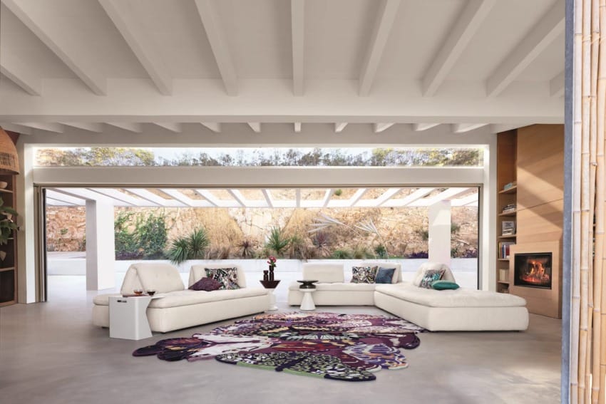 Spacious living room with white sofas, colorful rug, large windows, and modern fireplace, overlooking outdoor greenery.