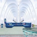 Futuristic white room with high arches, blue sectional sofa, colorful pillows, unique side tables, and a modern glass table.