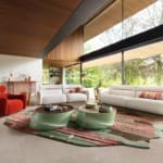 Modern living room with large windows, white sofas, red chairs, green coffee tables, and a nature view outside.