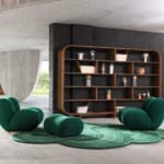 Modern lounge with green chairs, round rug, and wooden bookshelf against a black brick wall in a spacious, stylish room.