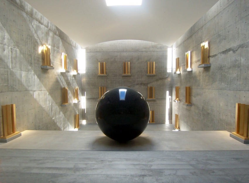 Modern concrete gallery space with large black sphere centerpiece, surrounded by gold-accented exhibits and natural light.