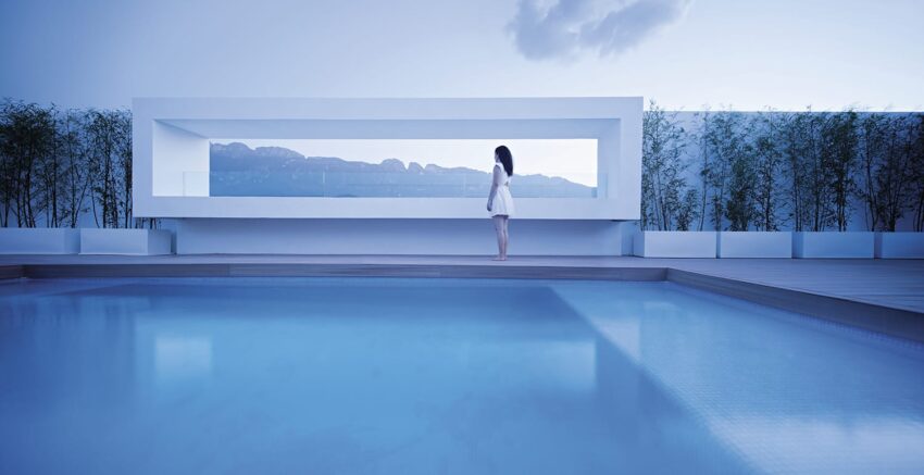 Person standing by a modern pool with a geometric structure in the background, surrounded by greenery and a serene sky.