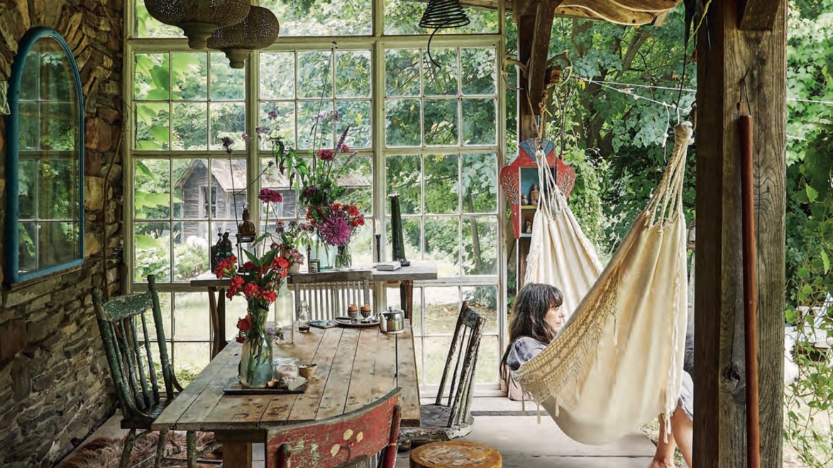 Cozy rustic room with hammock, wooden table, and chairs, surrounded by large windows and lush greenery outside.