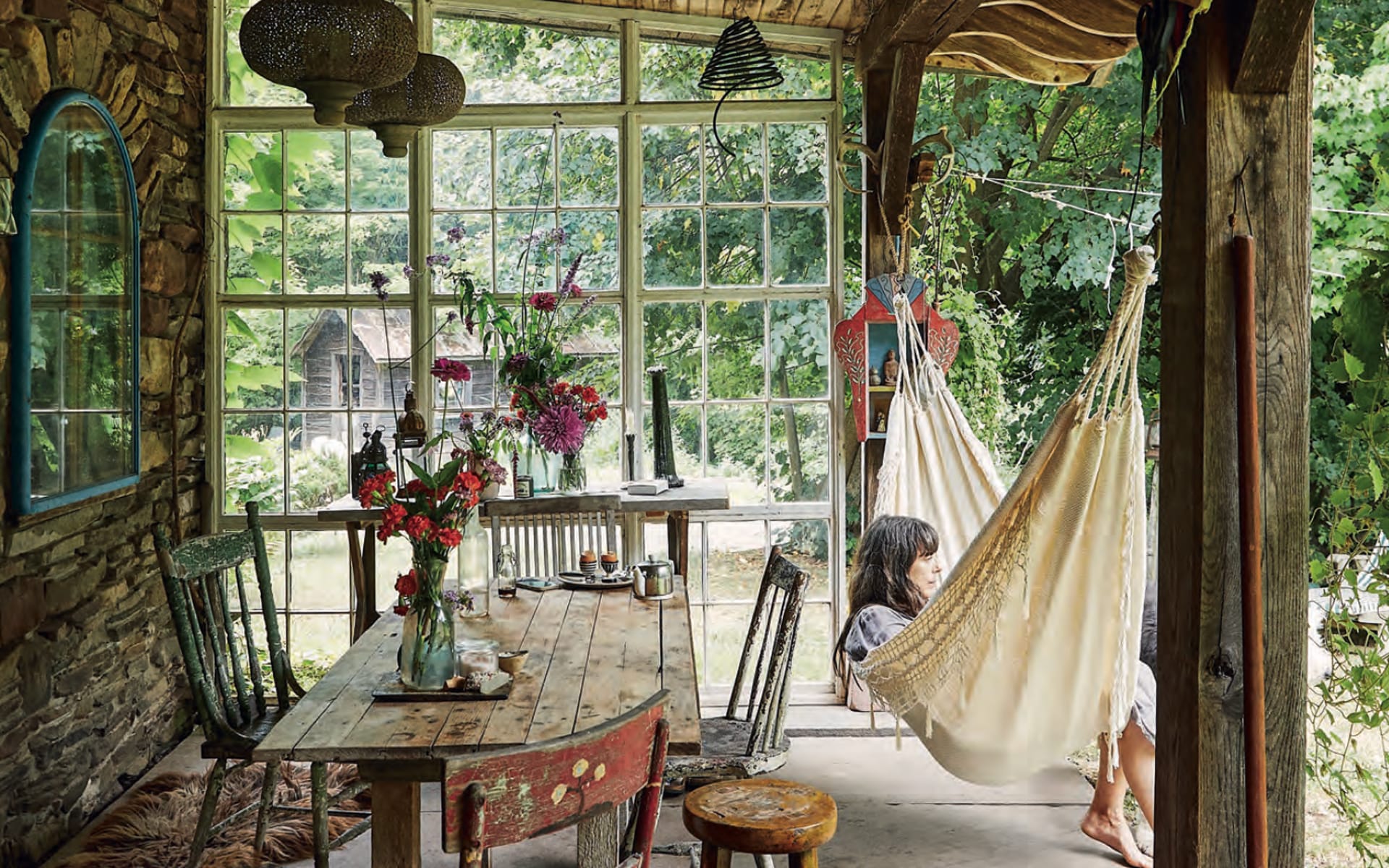 Cozy rustic room with hammock, wooden table, and chairs, surrounded by large windows and lush greenery outside.