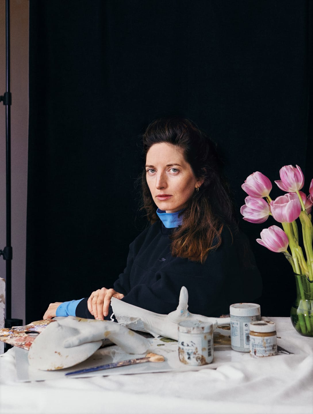 Woman seated at a table with art supplies and pink tulips in a vase, set against a black background.