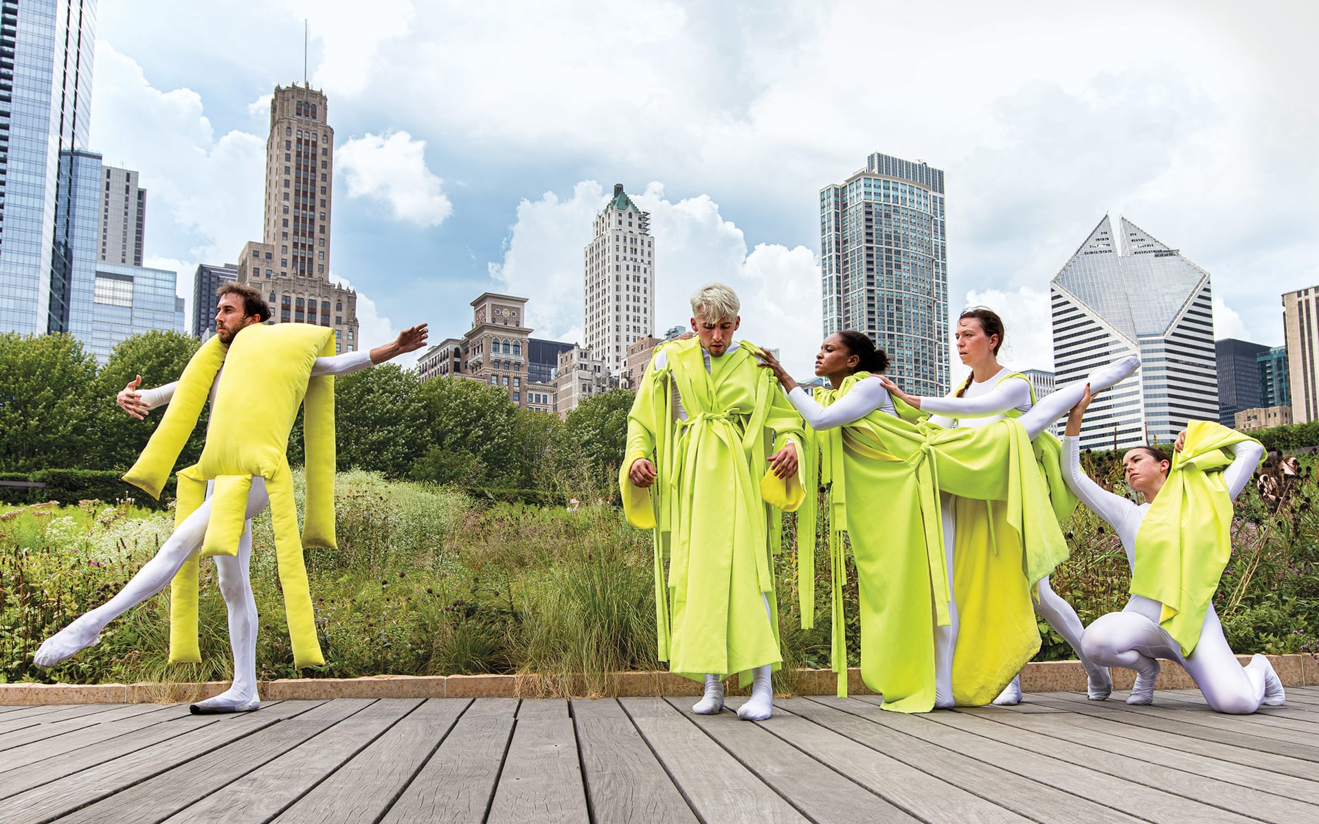 Dancers in bright yellow costumes perform a modern dance on a wooden platform in an urban park with skyscrapers in the background.