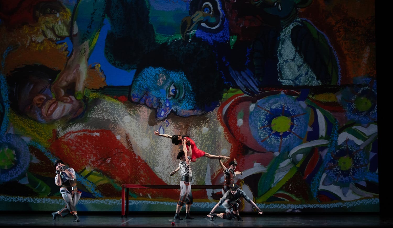 Dancers perform on stage in front of a vibrant, abstract mural featuring colorful figures and shapes.