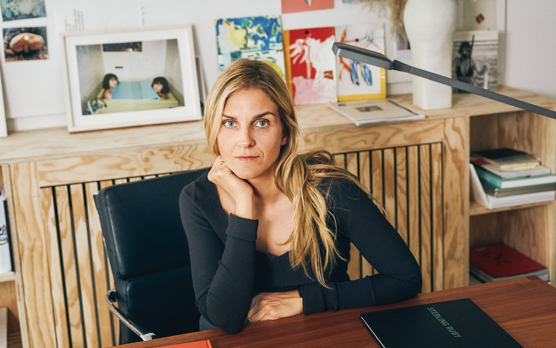 Person seated at desk in office, wearing a dark top, with hand resting on chin and shelves full of books and art in background