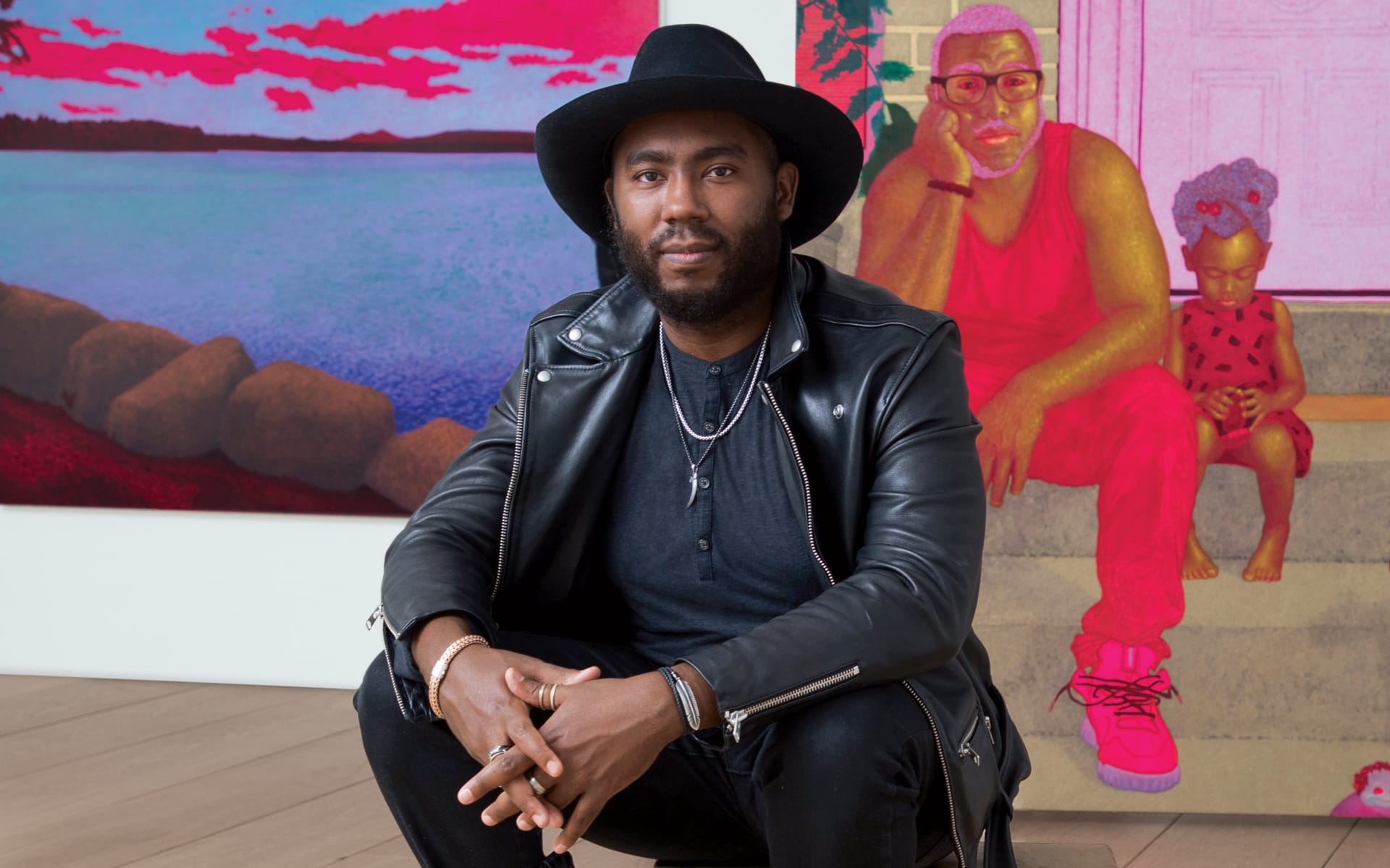 Person in black hat and leather jacket seated in front of colorful artwork with two figures in the background.