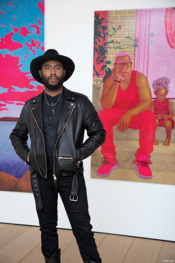 Man in black leather jacket and hat standing in front of colorful artwork depicting a person sitting with a child.