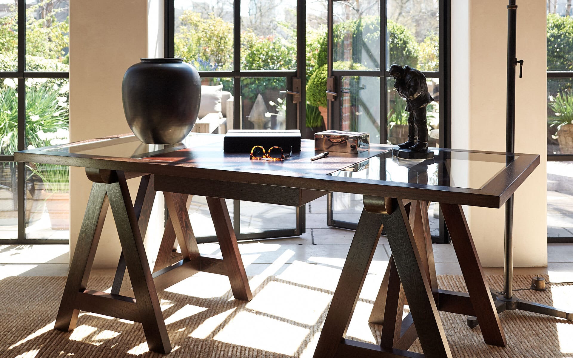 Elegant home office with a wooden desk, decorative vase, and sculpture, overlooking a garden through large windows.