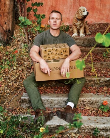Man sitting on outdoor steps holding boxes with a dog sitting behind him, surrounded by plants and a red wall.