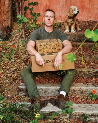 Man sitting on outdoor steps holding boxes with a dog sitting behind him, surrounded by plants and a red wall.