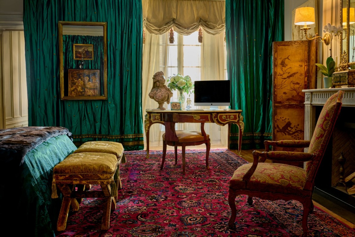 Elegant room with ornate furniture, green curtains, large rug, desk with flowers and bust, and a cozy fireplace.