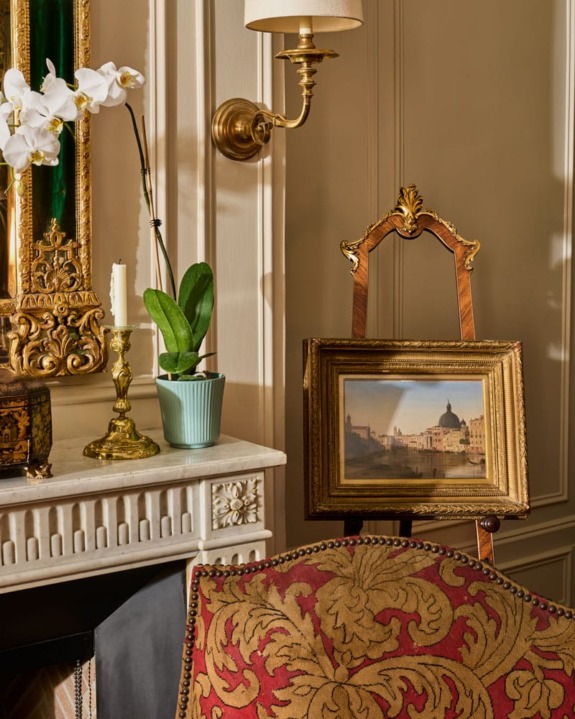 Elegant interior with ornate mirror, framed painting, orchid in pot, and upholstered chair near fireplace.