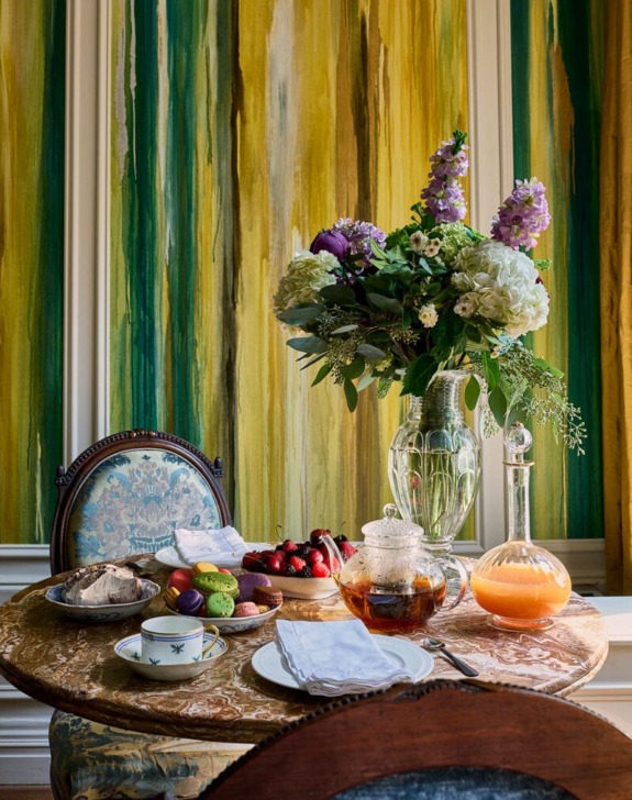 Tea set on a wooden table with flowers, fruits, and colorful macarons in front of a vibrant green and yellow striped wall.
