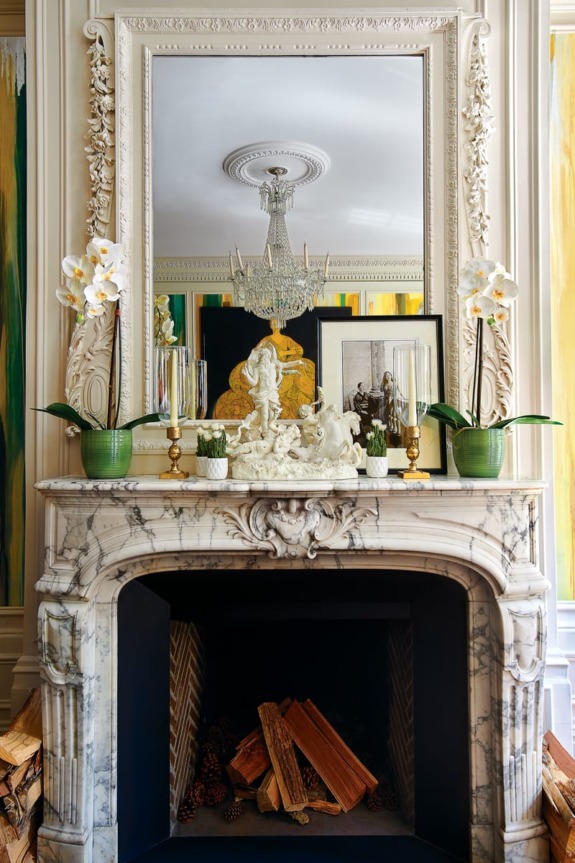 Marble fireplace with ornate mirror, decorative items, orchids, and firewood neatly arranged in the hearth.
