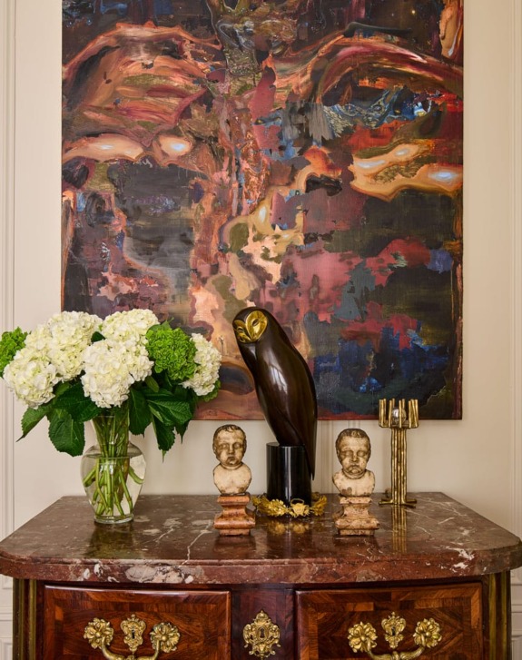 Marble dresser with owl sculpture, two busts, hydrangeas in a vase, and a large abstract painting in the background.