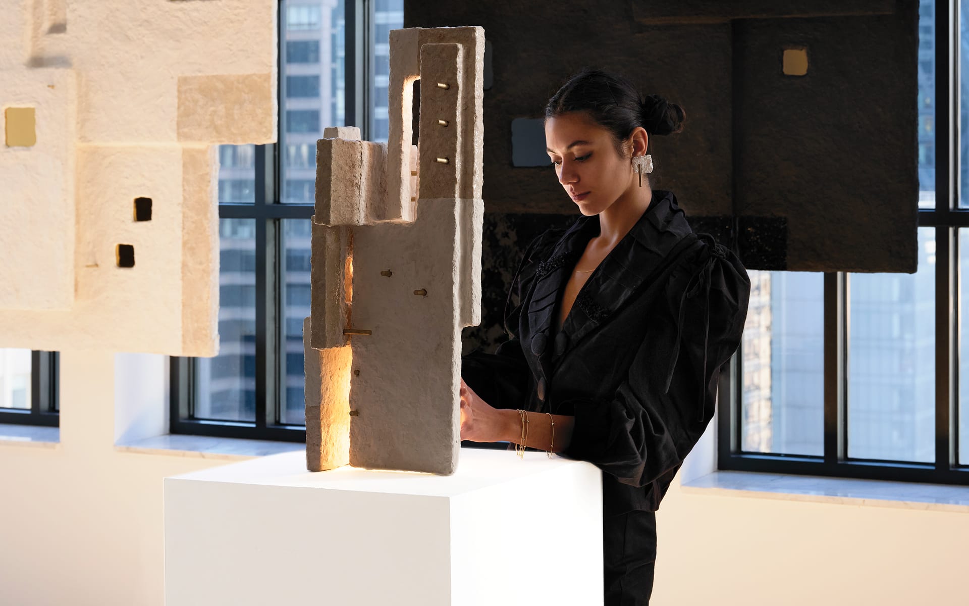 Person in black observing abstract sculpture on a pedestal in a gallery with large windows in the background.