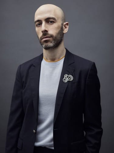 Man in dark suit jacket and light sweater, standing against a gray background, wearing a decorative pin on the lapel.