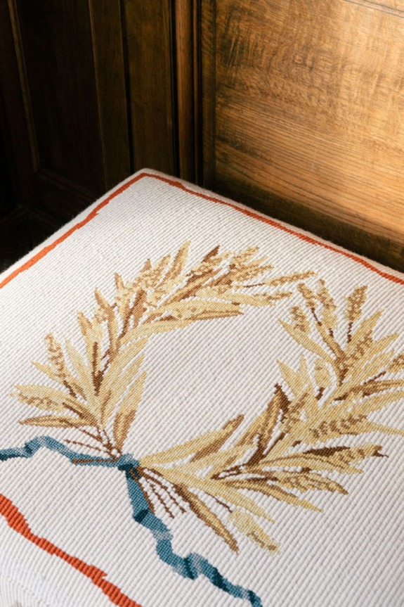 Cushion with embroidered wheat design and blue ribbon on wooden chair background.
