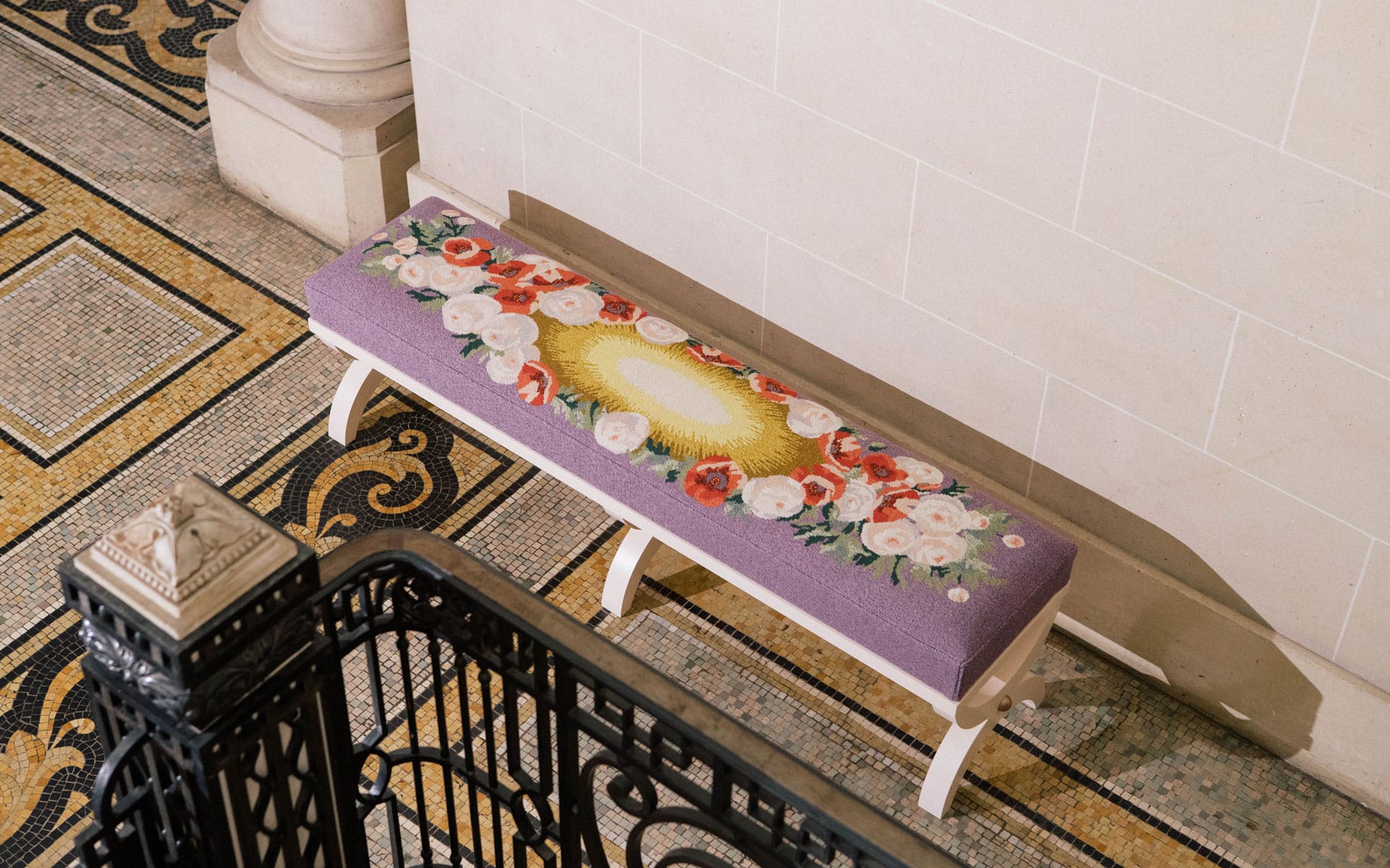 Purple bench with floral embroidery on tiled floor near a decorative staircase and beige wall.