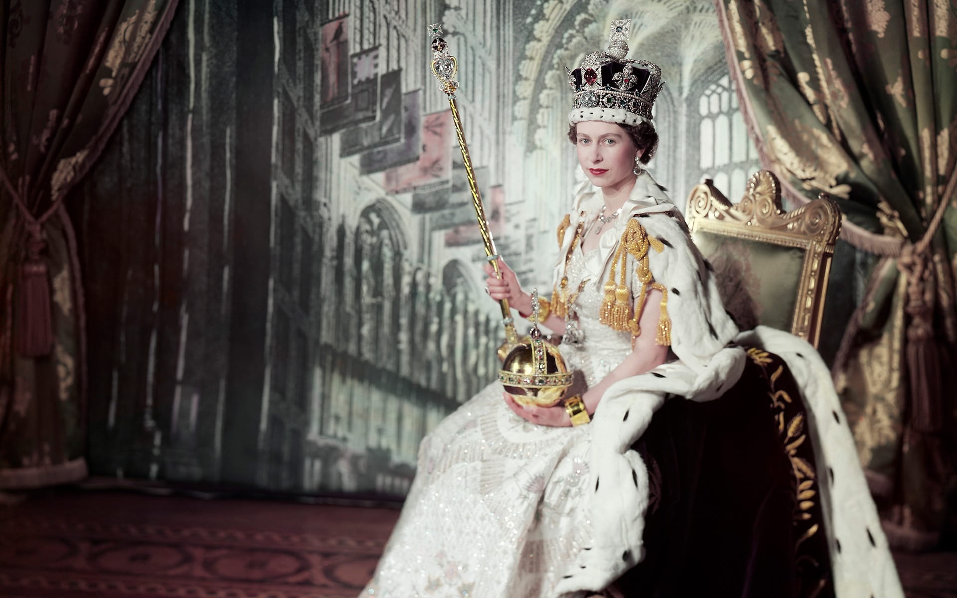 Woman in regal attire with crown and scepter, seated on a throne, in front of ornate backdrop.