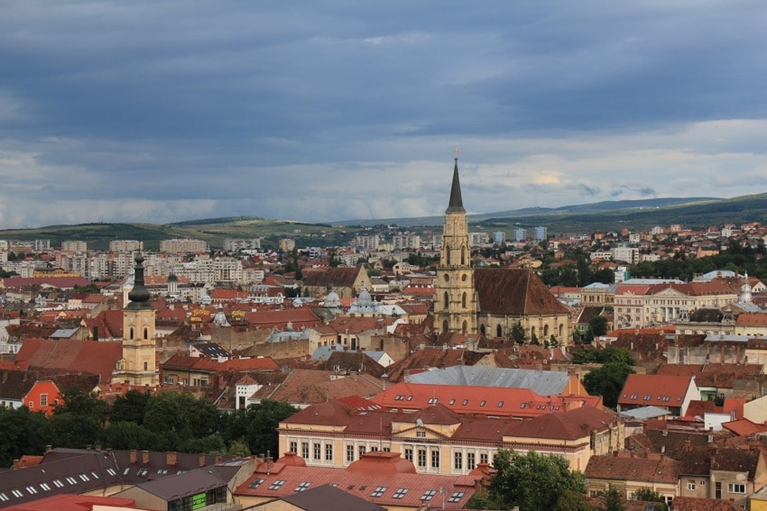 Aerial view of a historic European city with churches, red-roofed buildings, and a cloudy sky.