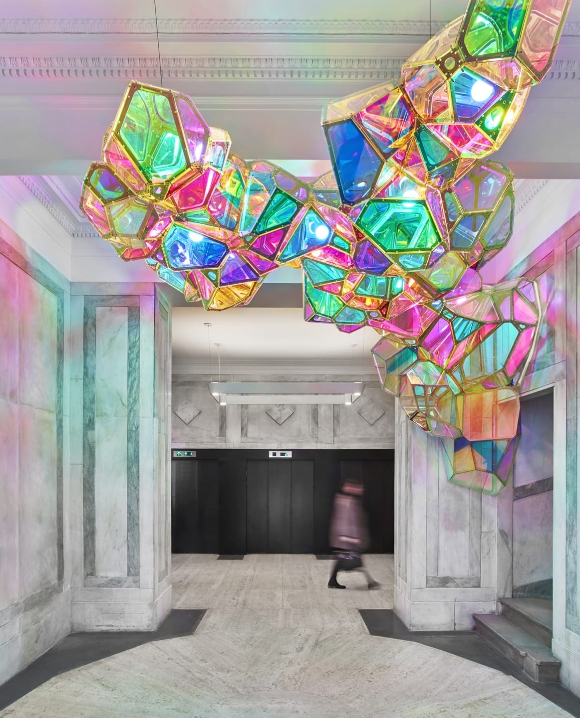 Colorful geometric light art installation in a modern building lobby with marble walls and elevators in the background.