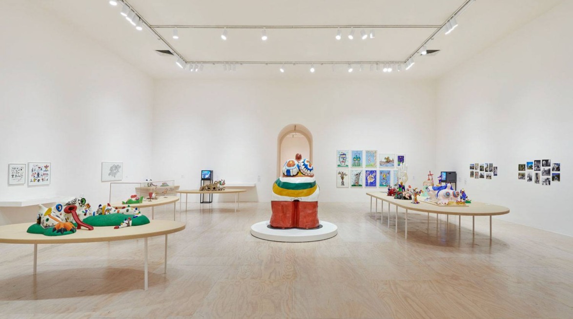 Contemporary art exhibition in a white-walled gallery featuring colorful sculptures and artwork on display tables.