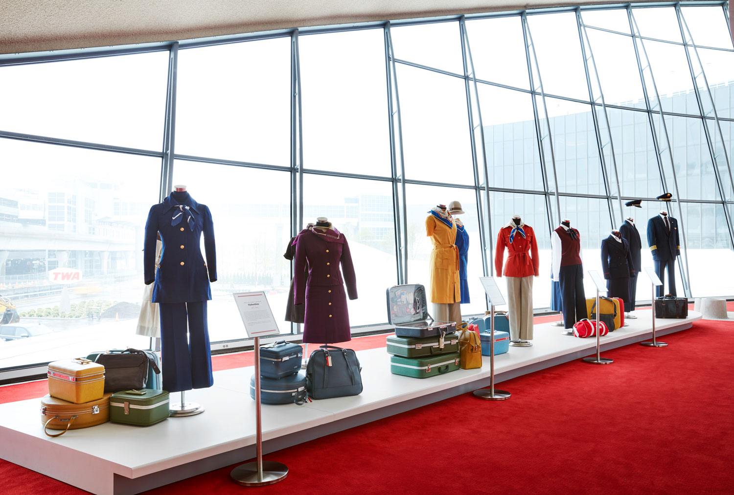 Mannequins displaying vintage airline uniforms on a platform with travel bags, set against large windows in a modern building.
