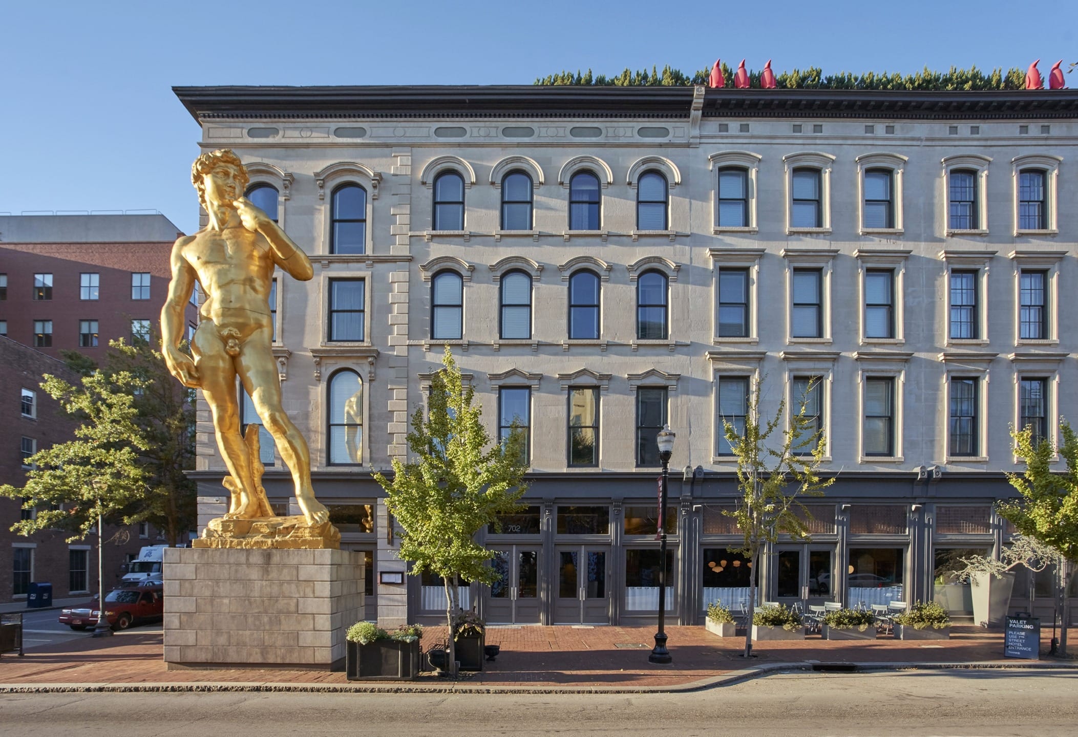 Golden statue of David replica in front of a historic building with arched windows and trees lining the street.