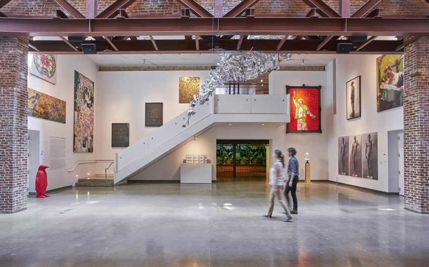 Art gallery interior with colorful artworks on the walls, a floating sculpture above, and people walking on a concrete floor.