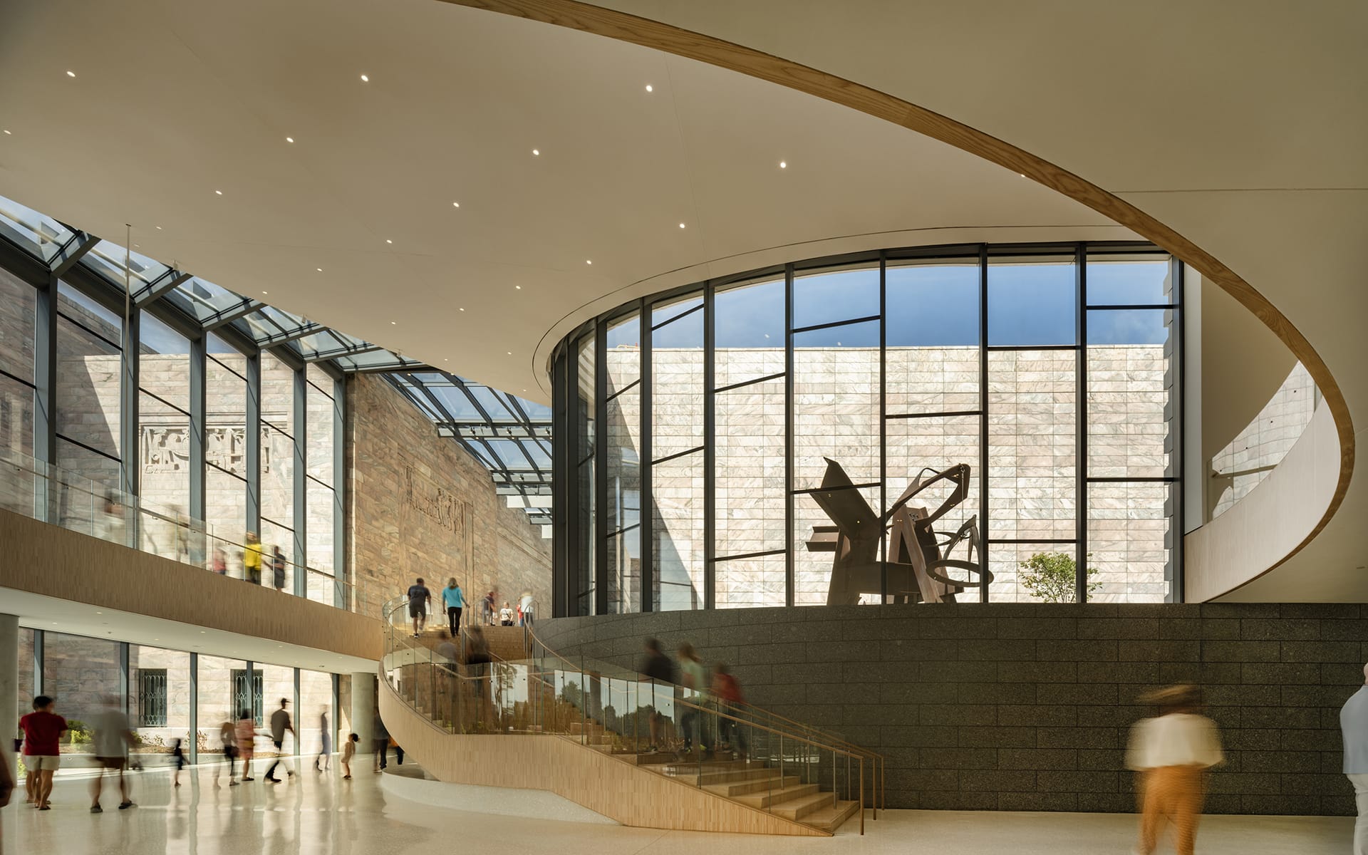 Modern museum interior with large windows, natural light, and abstract sculpture. People walking around the spacious area.