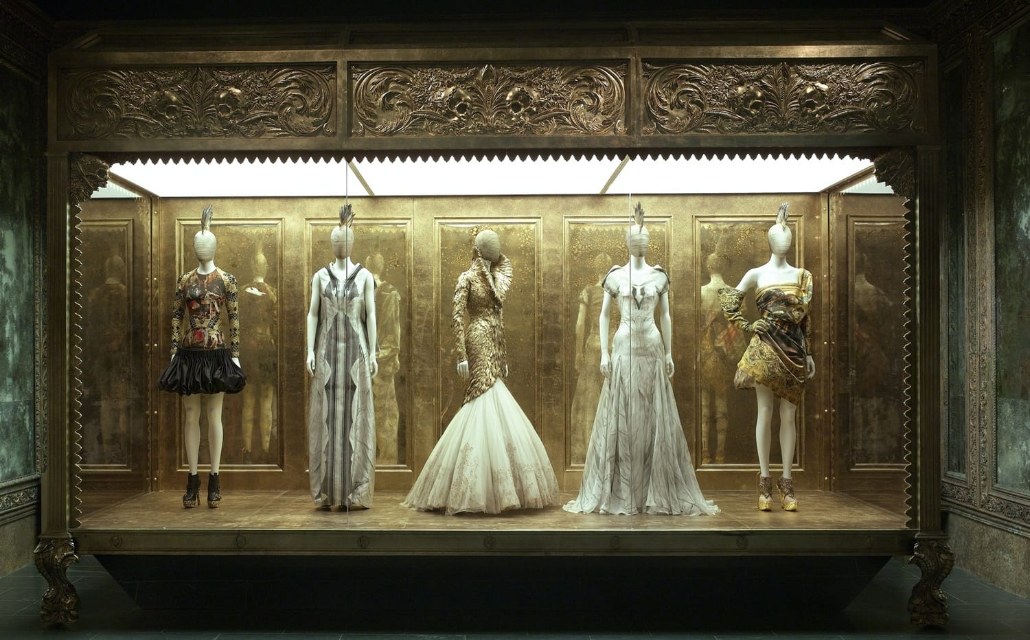 Fashion exhibit featuring five mannequins in ornate designer dresses displayed in an elegant, gold-adorned window setting.