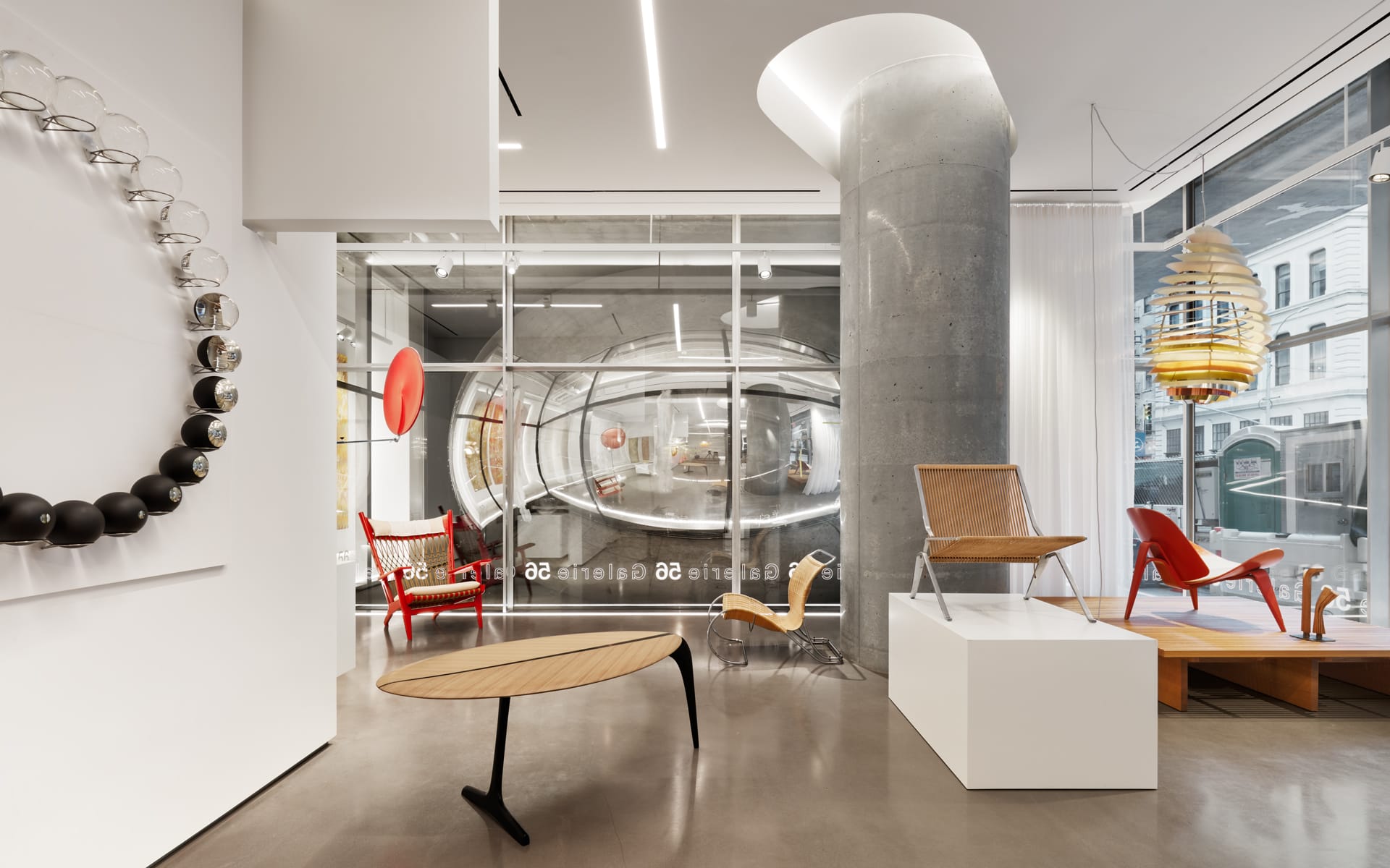 Modern furniture showroom with various chairs, tables, and decor against a glass wall and concrete column.
