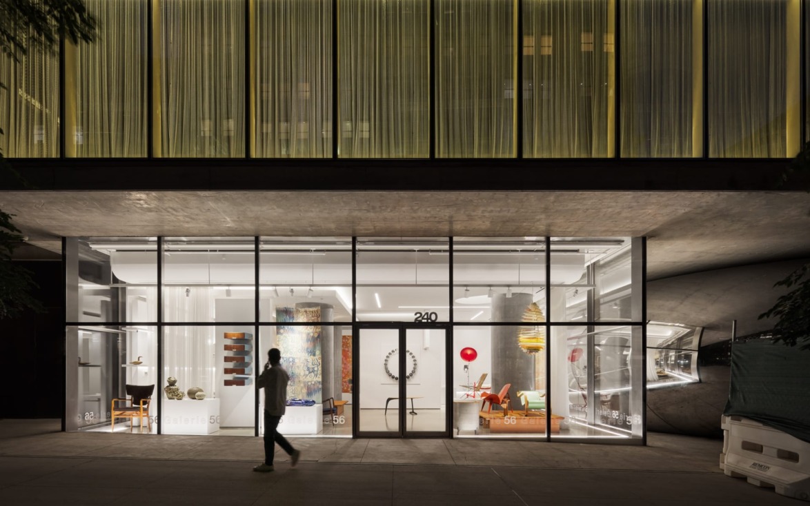 Street view of a modern building with large glass windows displaying furniture and art, person walking by at night.
