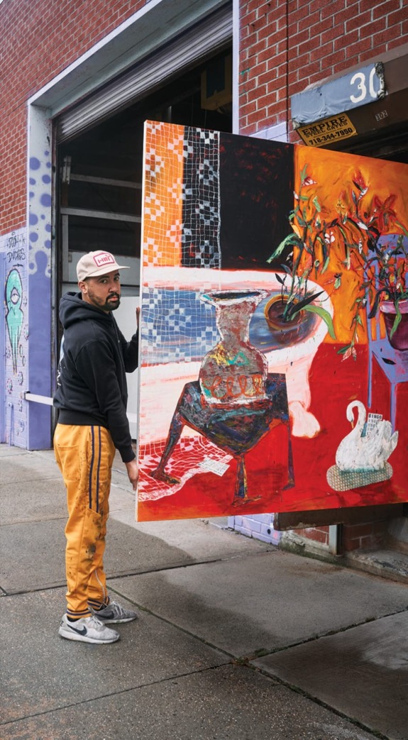 Artist moving a large, colorful painting outside a brick building with graffiti on the wall and an open garage door.