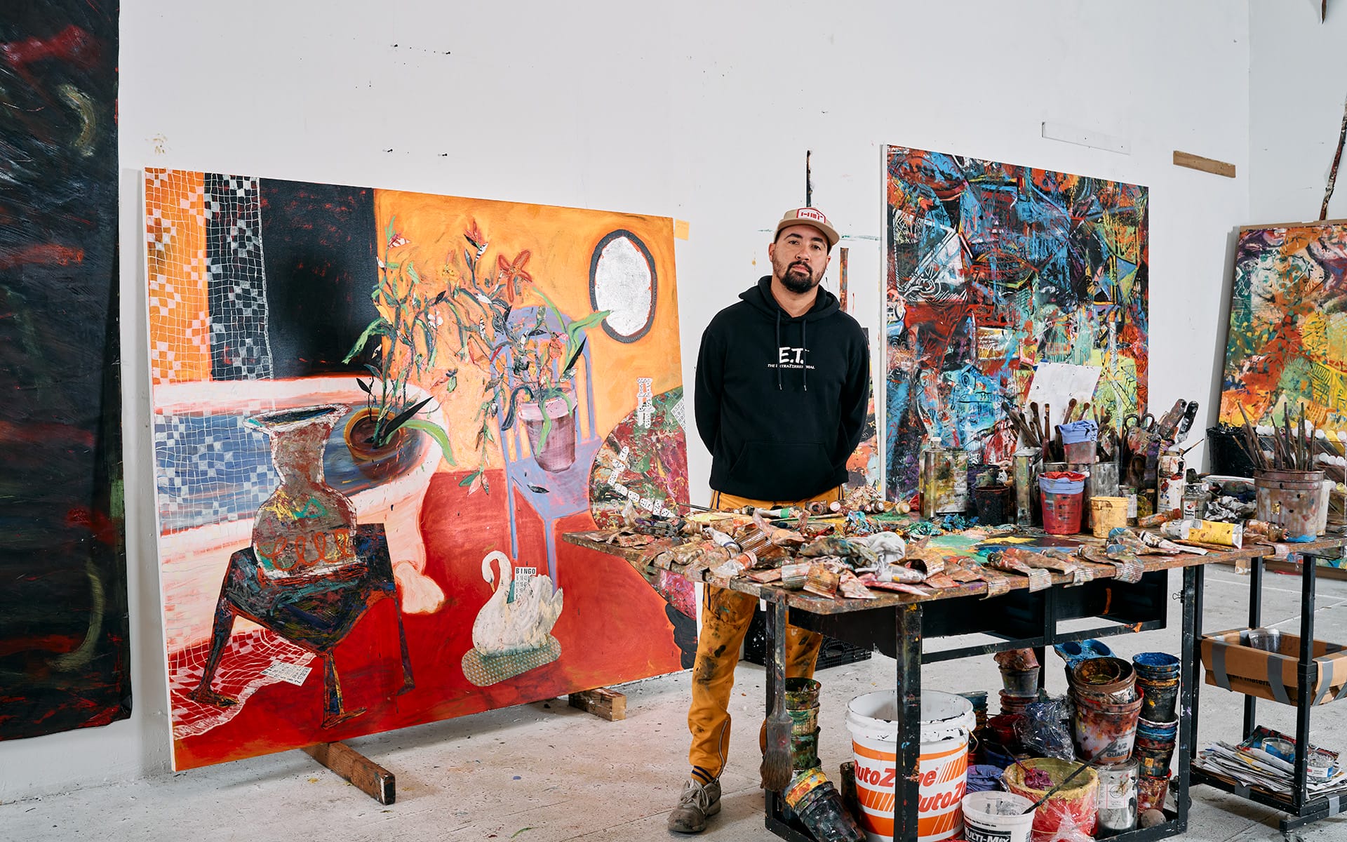 Artist standing in a colorful studio with vibrant abstract paintings and art supplies on a table.